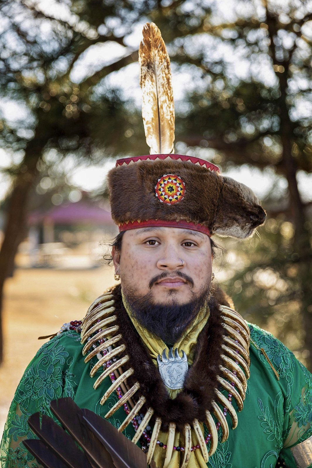 A man in a feathered headdress and beaded regalia looks into the camera, photographed outdoors in soft light.