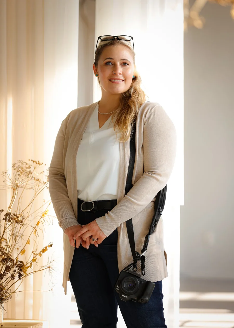 Janelle Nightingale standing in soft window light in a linen cardigan with a Canon camera hanging at her side.