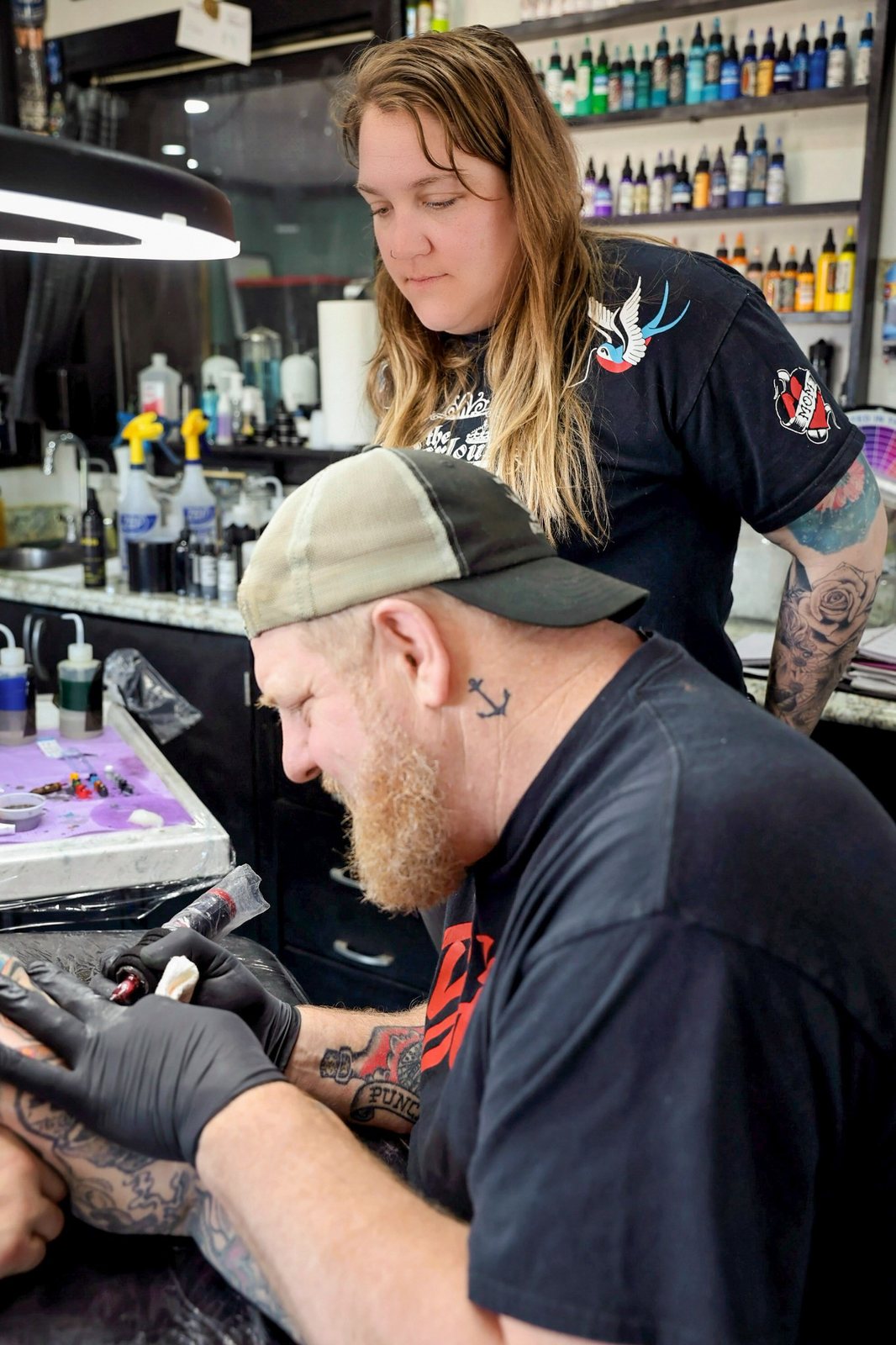 A tattoo artist in a ball cap bends over a client's arm at Stop the Hate in the 918, the free annual event in Sand Springs, Oklahoma, where artists cover hate and gang tattoos.