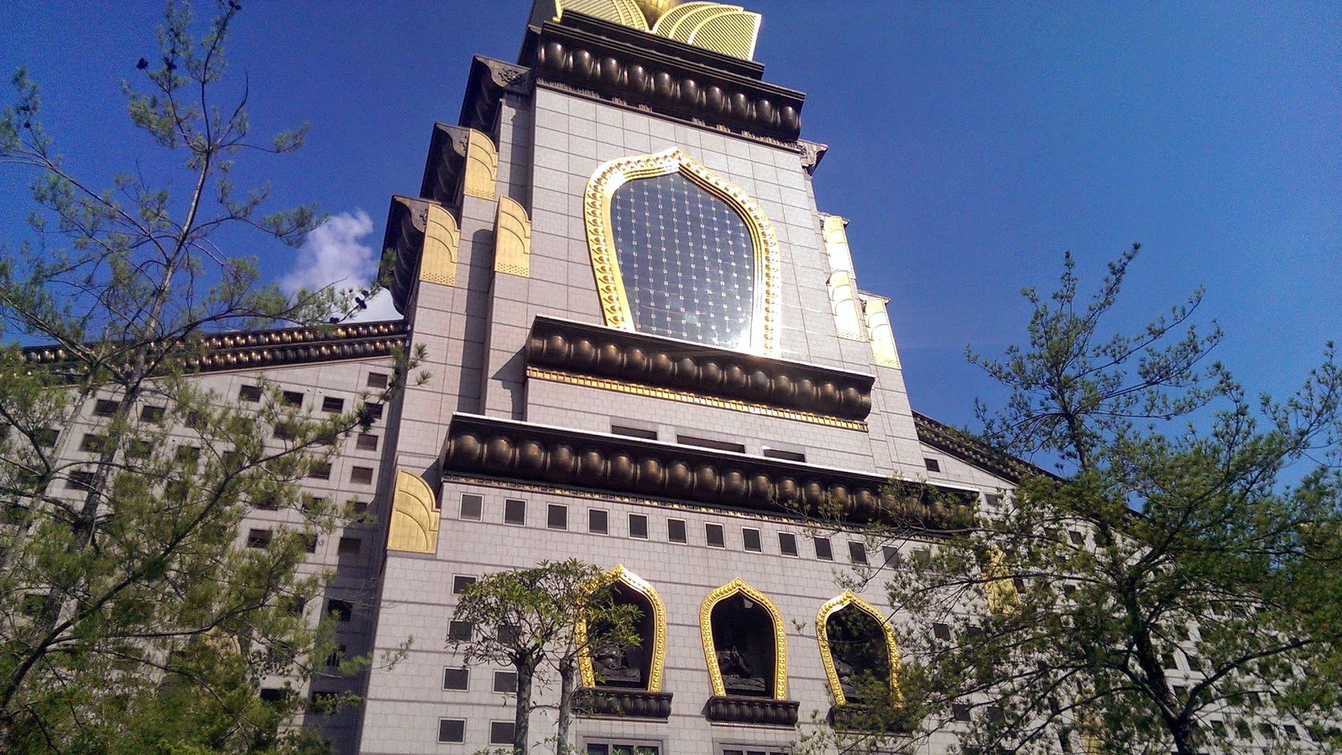 Looking up at the main tower of Chung Tai Chan Monastery in Puli, Taiwan, its gold trim bright against a deep blue sky and framed by pine branches.