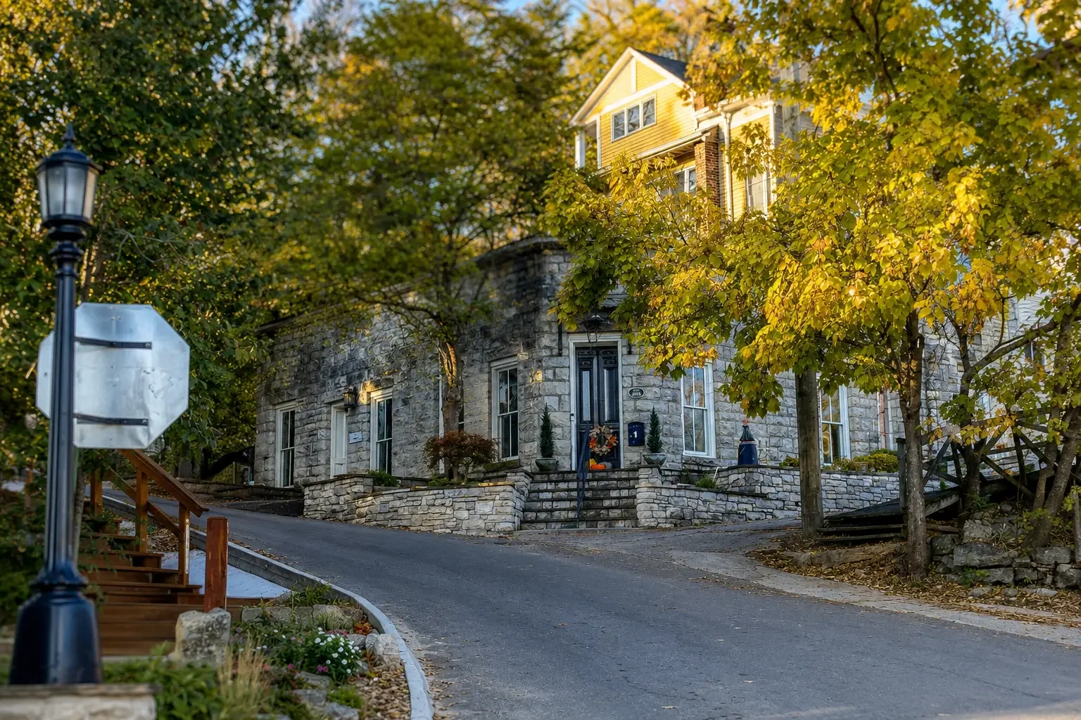 A sunlit small-town street with a stone building, trees, and a stop sign