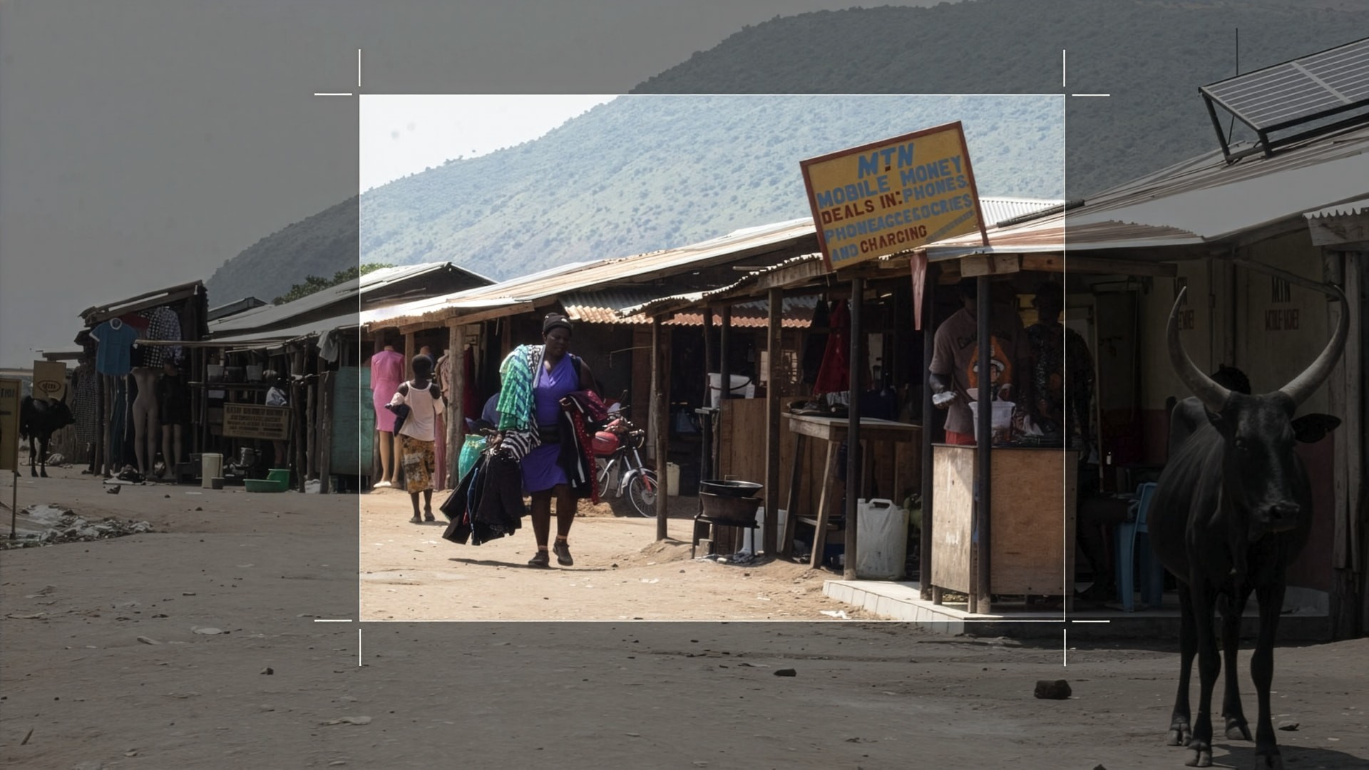A market street in rural Uganda, dimmed except for one framed section: a woman in a purple dress carrying an armload of clothing past small shops and a hand-painted MTN Mobile Money sign. A long-horned Ankole cow stands in shadow just outside the frame.