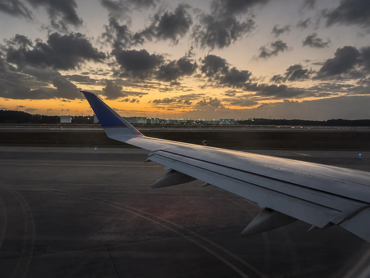 Airplane wing on a runway at sunset, viewed from a passenger window.